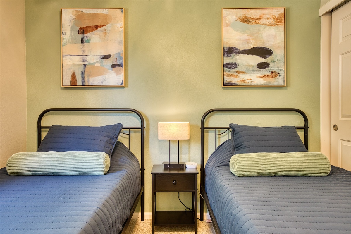 El dormitorio doble de color tranquilo incluye colcha suave, almohadas, mesita de noche, lámpara, estación de carga y cortinas traseras