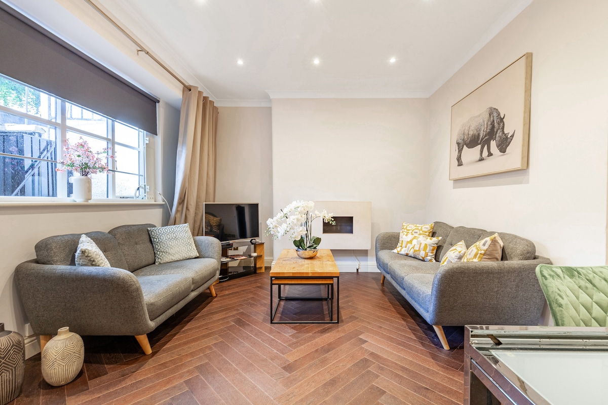 The living room features two cozy sofas with decorative cushions surrounding a wooden coffee table. A flat-screen TV is positioned on a stand, and natural light enters through a window adorned with simple curtains. The herringbone-patterned floor adds warmth to the space.