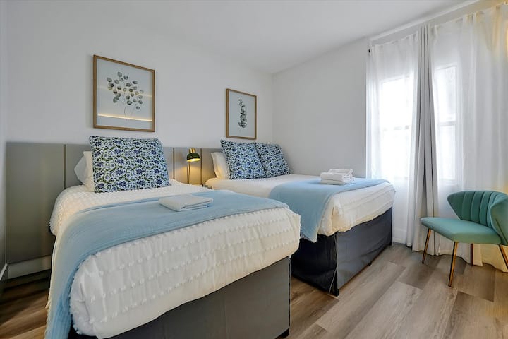 Chumba cha kulala chenye mwanga na kitanda cha watu wawili na kitanda cha mapacha, chenye madoido ya bluu laini na bafu la kujitegemea.