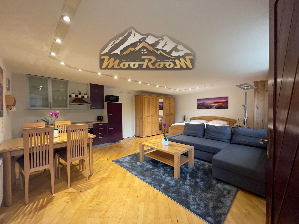 An open living area features a comfortable sofa and a wooden coffee table on a soft rug. A dining table with chairs is positioned nearby. Light wood cabinetry and appliances complement the modern kitchen area, while soft lighting enhances the overall warmth of the space.