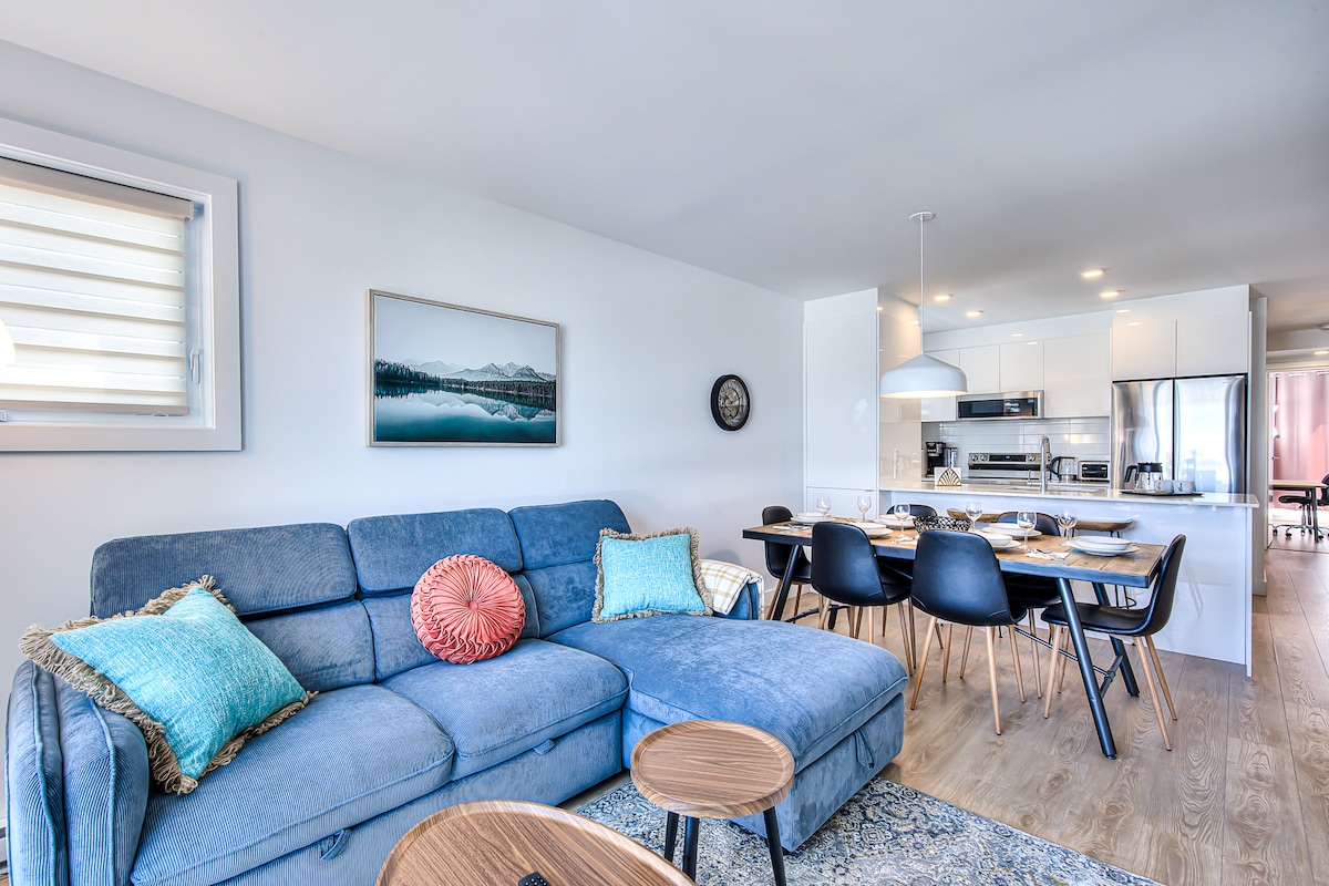 A modern living area features a comfortable blue sectional sofa complemented by decorative pillows. Adjacent, a dining table with black chairs is set for four, while a fully equipped kitchen is visible in the background, enhancing the space's inviting design.