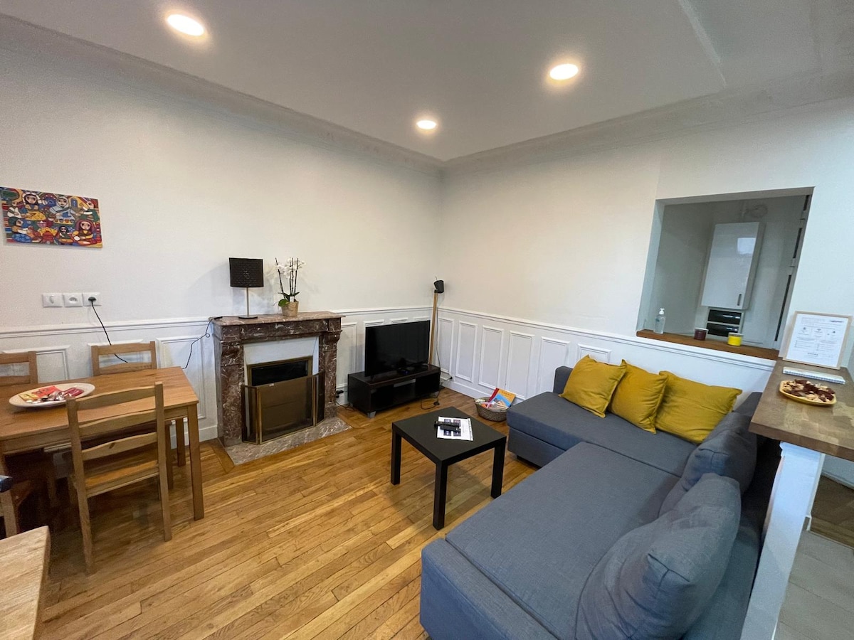 The living area features a gray sectional sofa with yellow cushions, accompanied by a rectangular black coffee table. A flat-screen television is positioned next to a decorative fireplace. A wooden dining table with chairs is visible, enhancing the cozy and functional space.