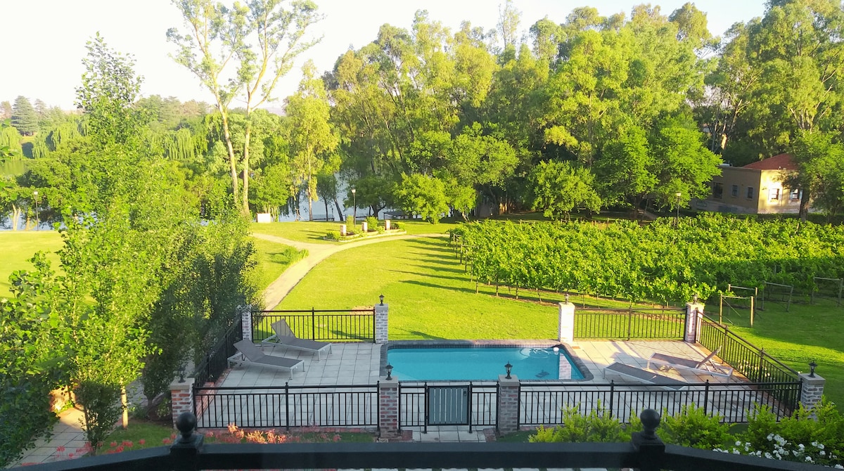 A tranquil view from a balcony showcases a private swimming pool surrounded by a stone deck. Lush greenery surrounds the area, with well-maintained landscaping leading towards the serene Vaal River. A vineyard can be seen in the distance, offering a sense of peace and natural beauty.