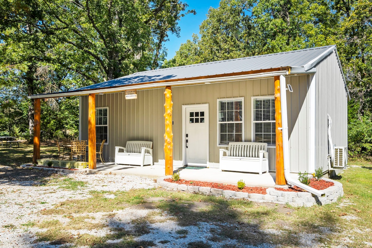 A cozy cabin exterior is presented with a light grey metal facade and wooden accents. Two benches are positioned on the porch, surrounded by neatly arranged landscaping. The entrance features a central door with a window above it, inviting guests to enjoy the outdoor space.