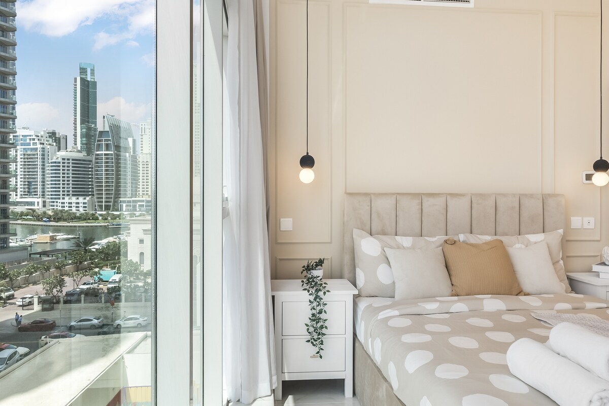 A bright and airy bedroom features a spacious bed adorned with a polka-dotted duvet and soft accent pillows. Large windows provide a stunning view of the vibrant city skyline, complemented by sheer curtains allowing natural light to fill the room.