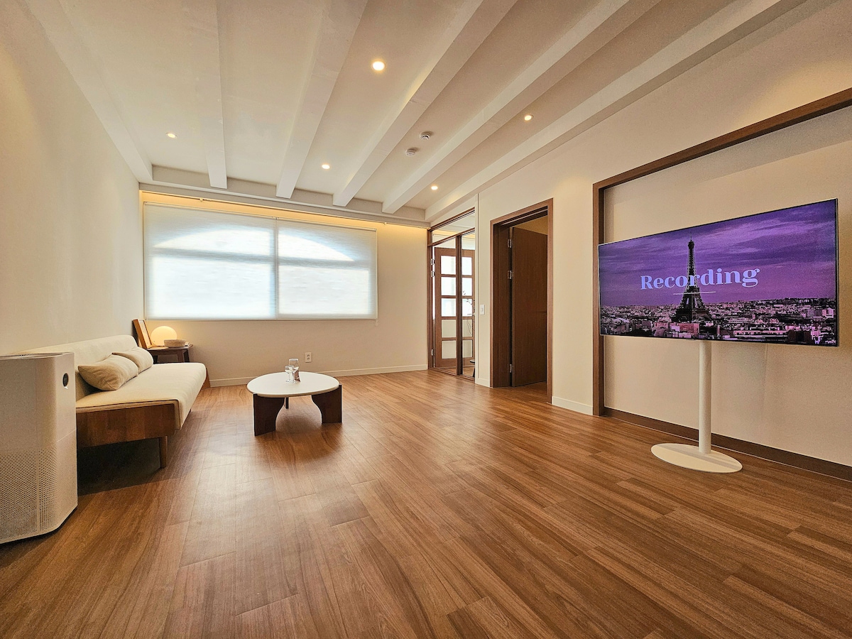 A spacious living room with warm wooden flooring features a comfortable seating area, including a cozy couch. A large window allows natural light to fill the space, while a flat-screen TV stands on a sleek pedestal, showcasing a scenic city view.