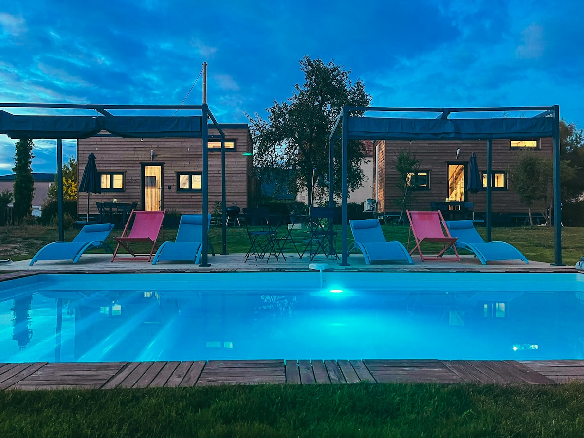 A tranquil view of a pool area is featured, with lounge chairs arranged under shaded canopies. Two tiny houses are visible in the background, surrounded by greenery. Soft lighting from the windows of the houses casts a gentle glow as dusk settles in.