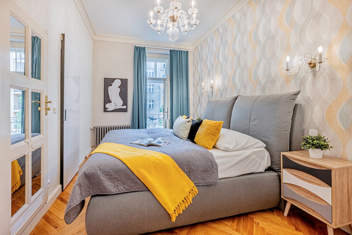 The spacious bedroom features a large, comfortable bed with a decorative yellow throw. Elegant wall lighting and a crystal chandelier enhance the ambiance. A mirror reflects the stylish design, while soft, cascading curtains frame the window, which opens to views of the outdoor surroundings.