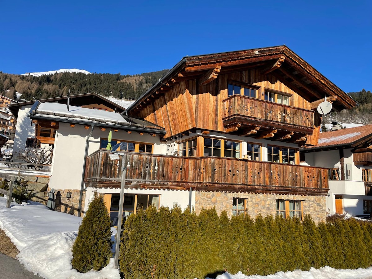 The chalet features a traditional wooden façade with balconies adorned with railings. Snow covers the ground in front, contrasting with the green hedges. A clear blue sky and mountain backdrop contribute to the serene environment, highlighting the chalet’s inviting appeal.