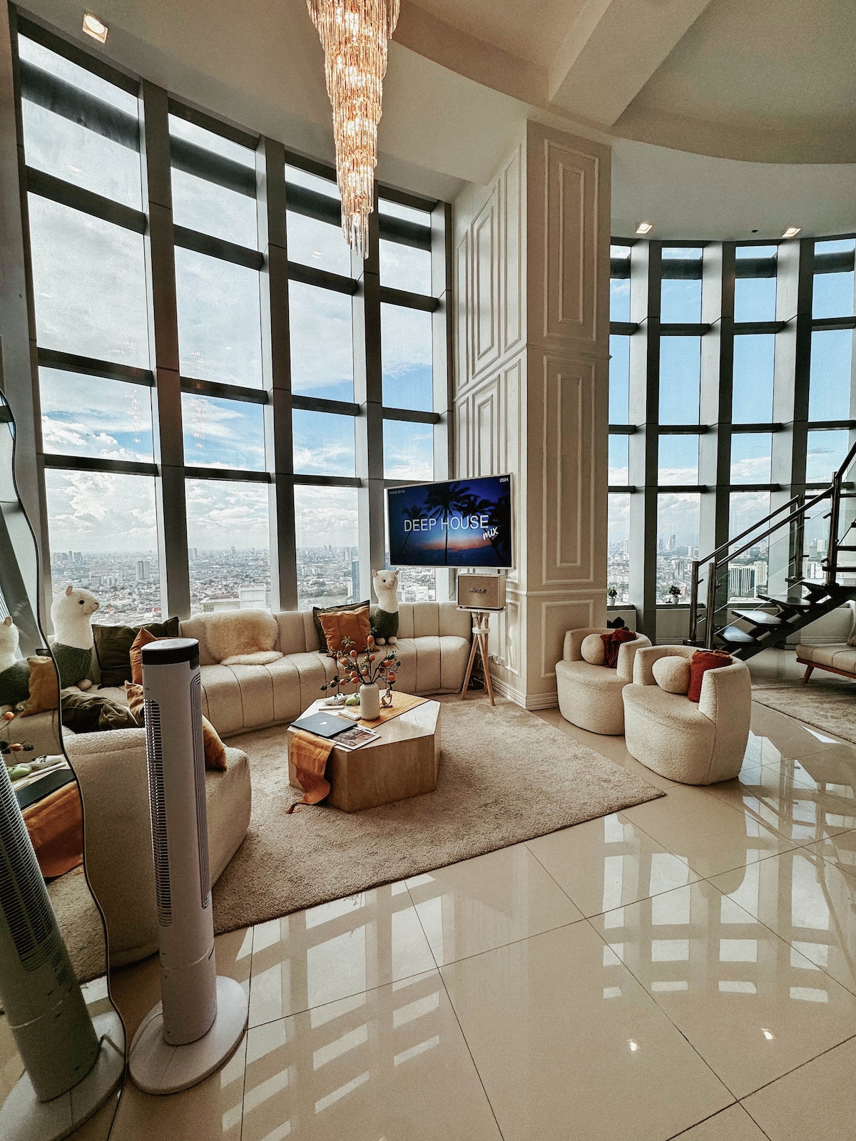 The living area features a circular layout with large windows providing panoramic city views. A comfortable seating arrangement includes plush chairs and a sofa surrounding a low coffee table. A sleek television is mounted on the wall, and light reflects off the polished floors, enhancing the bright atmosphere.