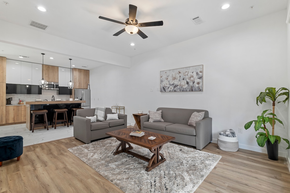A modern living area features two cozy sofas and a wooden coffee table. Natural light fills the space through large windows, with greenery added by a nearby plant. The open kitchen can be seen in the background, showcasing wooden cabinetry and contemporary appliances.