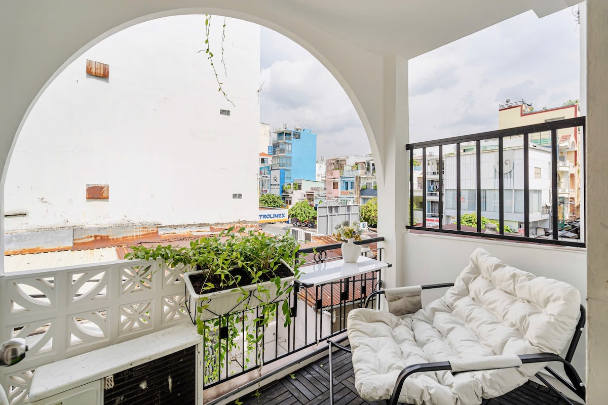 A private balcony area features a comfortable seating arrangement with a plush white chair and a small table. Lush greenery cascades from planters, complementing the urban view of nearby buildings and streets. The space is well-lit by natural light from the above arch.