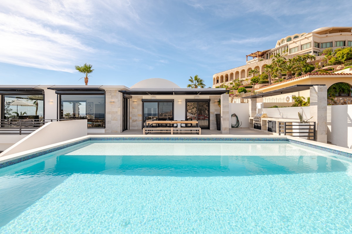 A spacious outdoor area is depicted, focusing on a shimmering pool that reflects the sky. An elegant seating area is visible beyond the pool, with large glass doors leading into the villa. Lush landscaping surrounds the space, enhancing the serene ambiance.