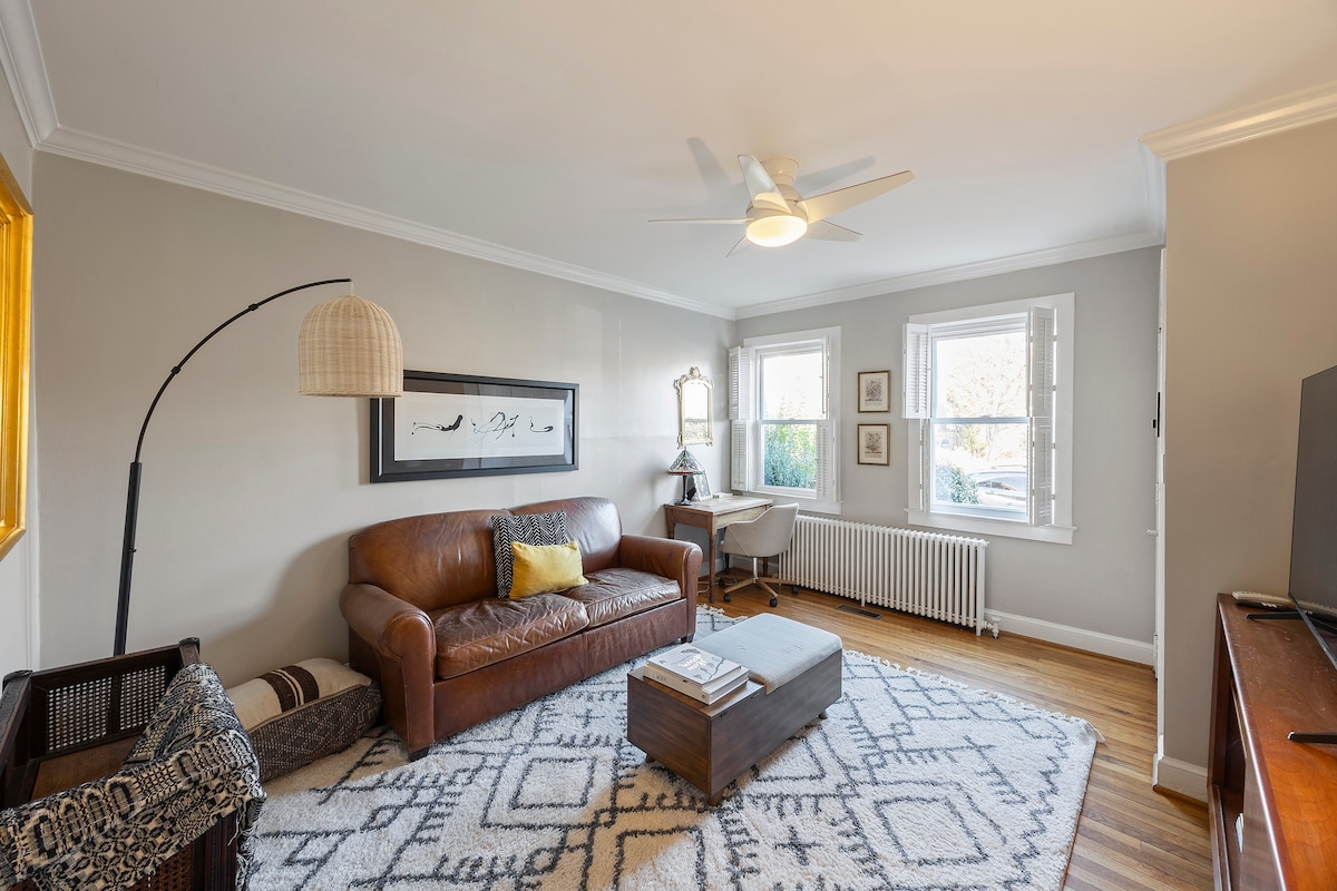 A welcoming living room features a brown leather sofa accompanied by a light-colored patterned area rug. Natural light enters through two side windows, illuminating the room. A ceiling fan and adjacent workspace provide functionality, while art is displayed on the walls, adding character.