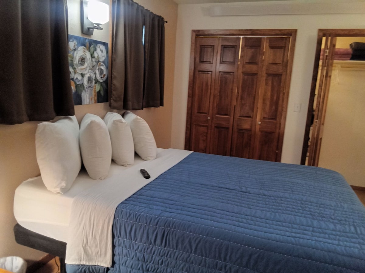 A comfortable sleeping area features a spacious bed dressed in light blue bedding, accentuated by several white pillows. A floral artwork hangs on the wall, and the room is illuminated by a wall-mounted light fixture. Dark curtains frame the window, and wooden double doors provide storage.