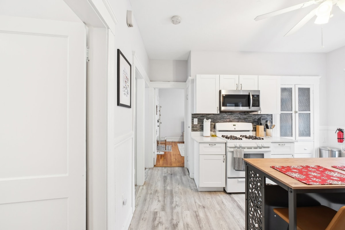 A bright kitchen area is equipped with modern appliances, including a gas range and microwave. Light-colored cabinetry provides ample storage, and a dining table with a decorative table runner adds a touch of color. Natural light fills the space through the windows, enhancing the airy atmosphere.