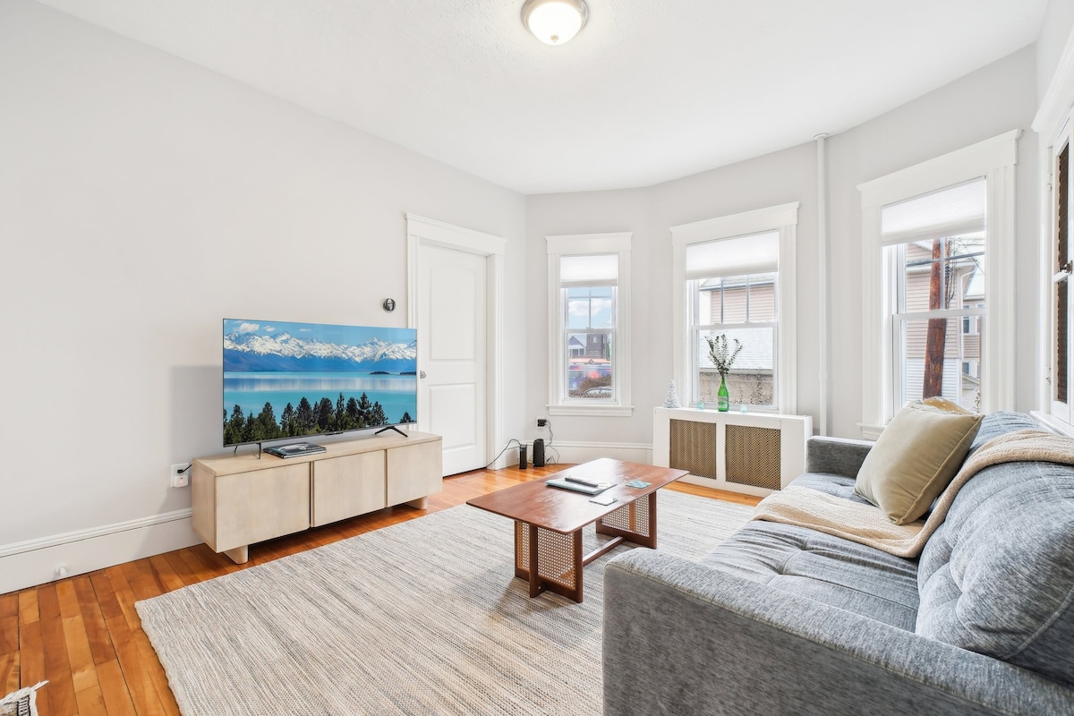 A spacious living area highlights a comfortable gray sofa, complemented by a wooden coffee table at the center. A large television is mounted on the wall, and natural light enters through multiple windows adorned with white trim.