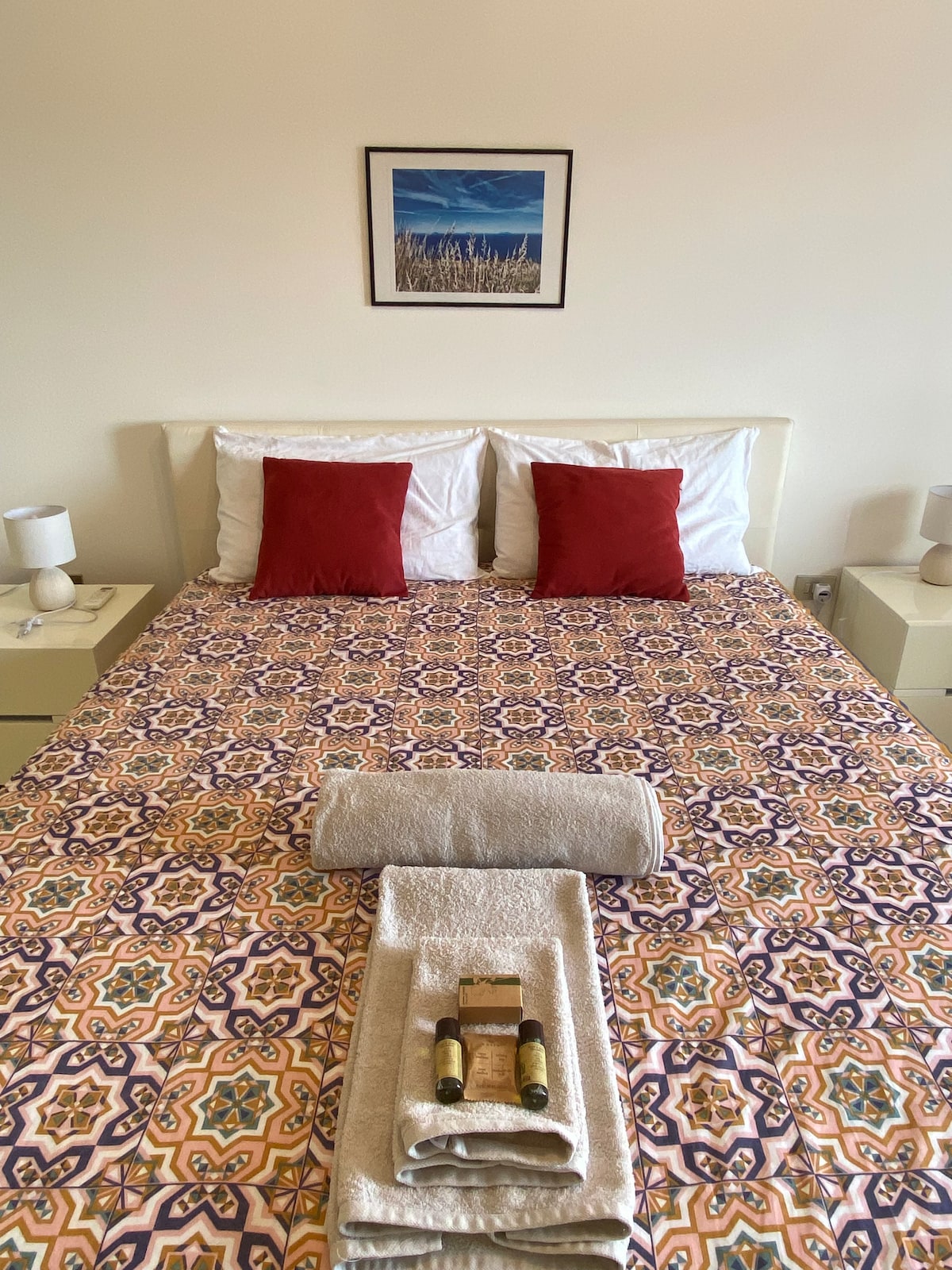 A spacious double bed is centered in the room, adorned with a patterned bedspread in warm tones. Two red accent pillows add color, while bedside tables with lamps are situated on either side. Neatly folded towels and toiletries are arranged at the foot of the bed.