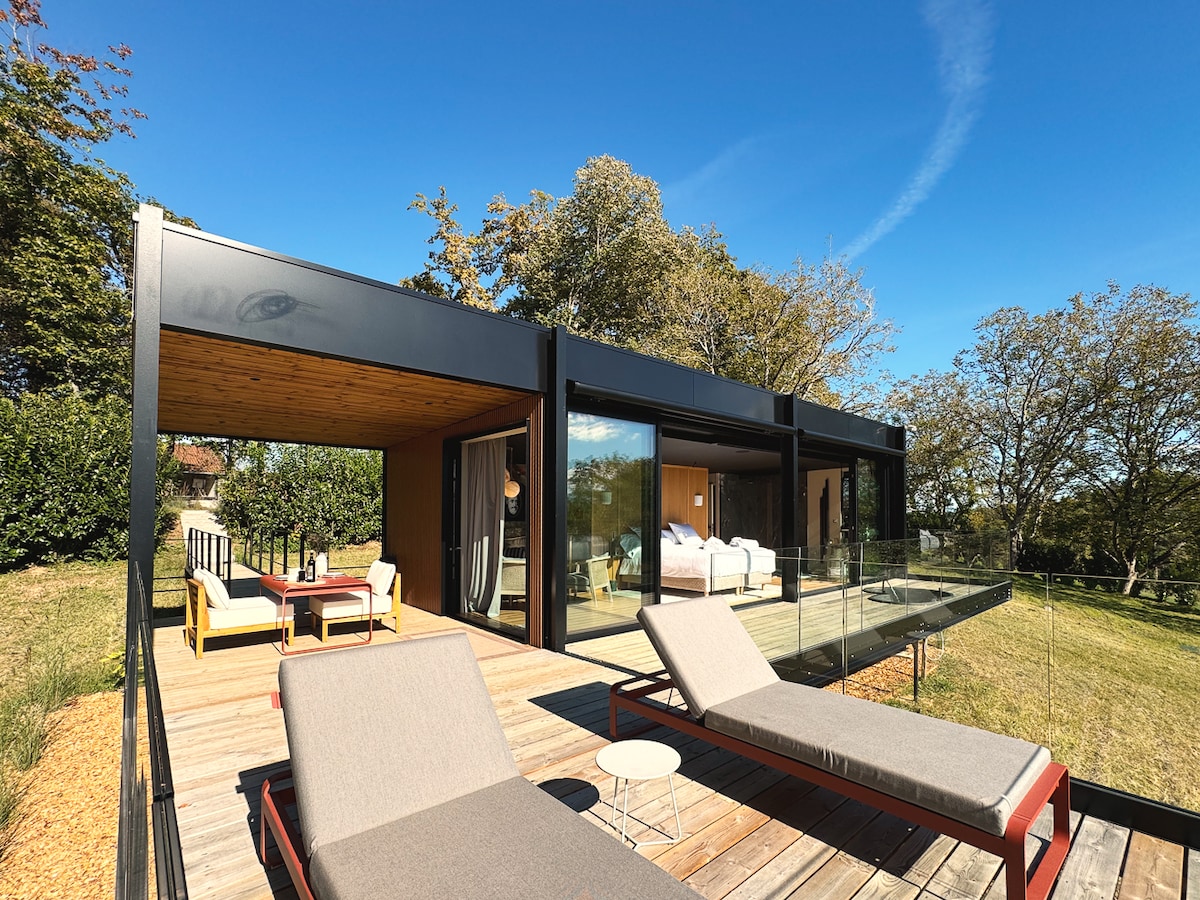 A modern suite is shown with large glass windows overlooking a lush landscape. A spacious deck features two lounge chairs and a small table, providing a serene outdoor seating area. The structure blends contemporary design with natural surroundings.