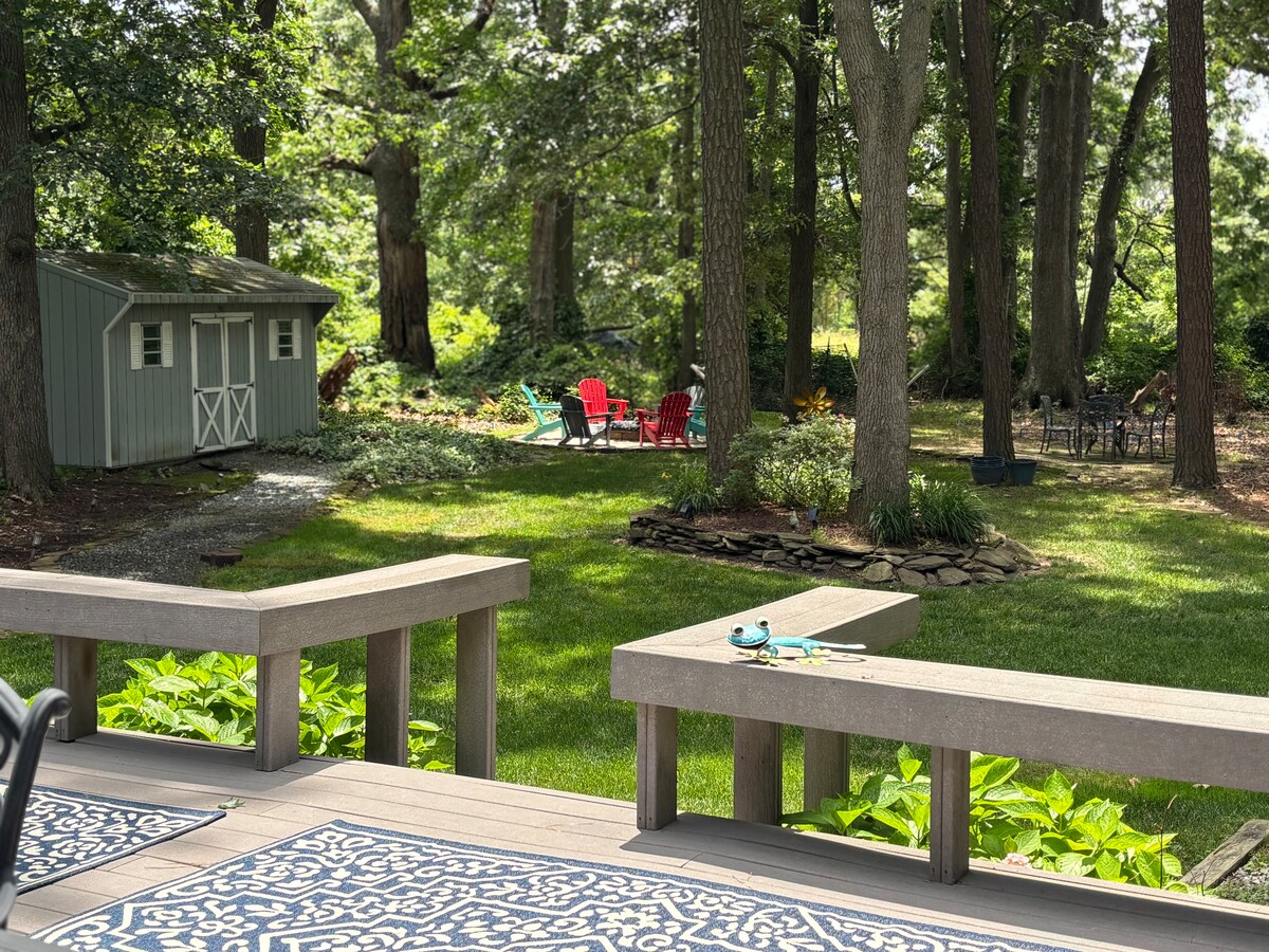 A spacious backyard is surrounded by tall trees, providing a serene setting. Colorful Adirondack chairs are arranged in the distance, complemented by a small storage shed. A well-maintained lawn features lush greenery and natural stone landscaping, suitable for outdoor gatherings.