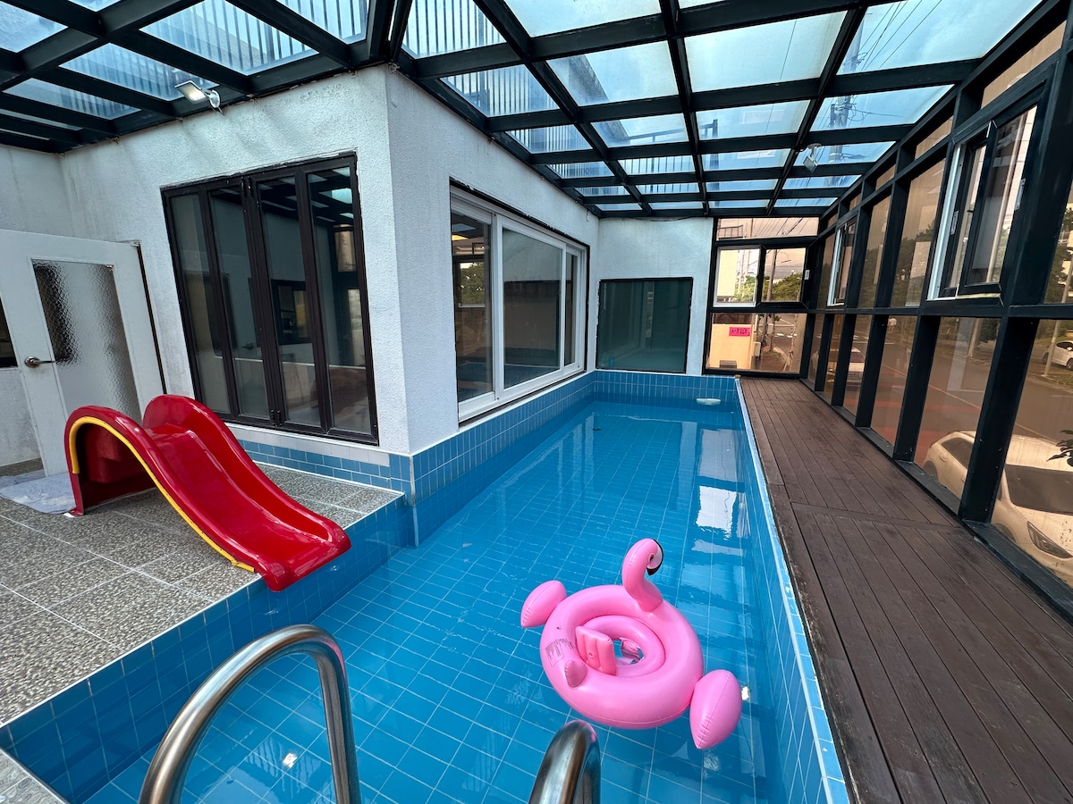 An indoor swimming pool features a bright blue tile surface, accompanied by a red slide and a pink inflatable float. Large windows allow natural light in, enhancing the space. Steps with a railing provide access to the pool area.