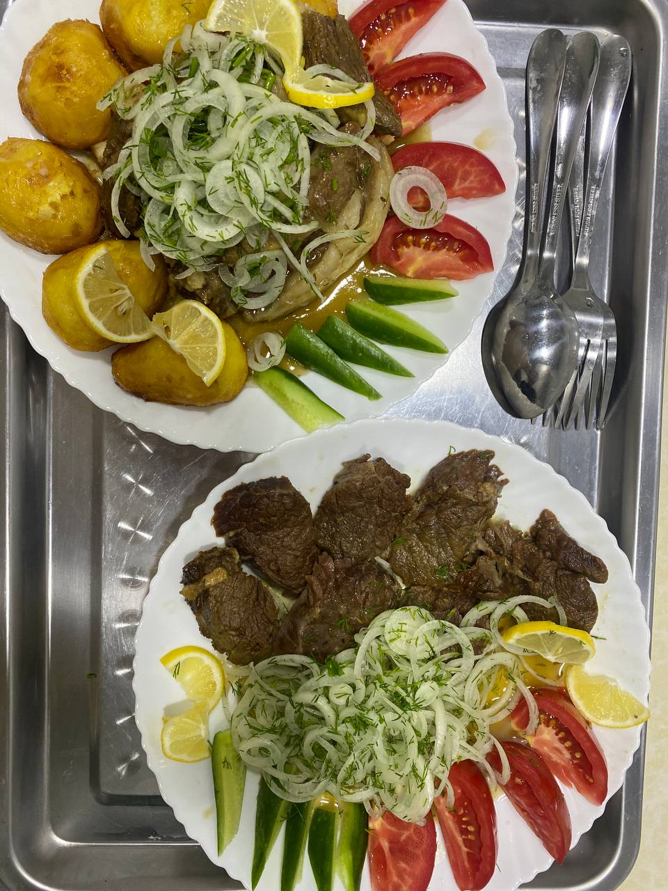 Two plated meals are presented on a silver serving tray. One plate features grilled meat, topped with chopped onions and herbs, accompanied by slices of tomato, cucumber, and lemon, alongside roasted potatoes. The second plate displays a similar arrangement without potatoes, emphasizing the meat and garnishes.