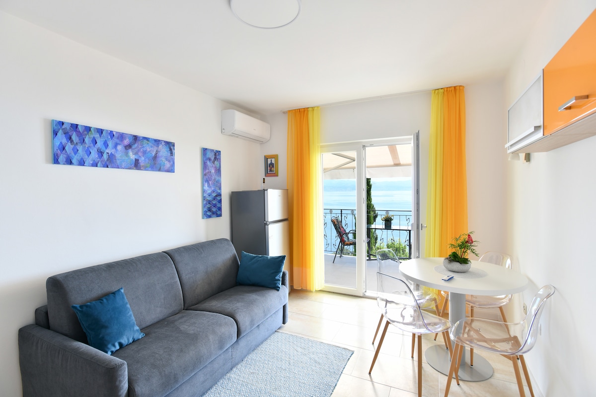 A bright living area features a gray sofa adorned with teal cushions and a small dining table with clear chairs. Large doors open to a balcony with a view, while pale yellow curtains add a touch of warmth. Modern artwork is displayed on the walls.