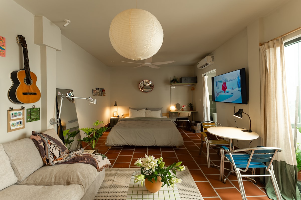 The studio features a spacious layout with a queen size bed, a comfortable sofa, and a small dining table. A 55-inch TV is mounted on the wall, and greenery is present throughout the room. Natural light enters through large windows, complemented by a lantern-style ceiling fixture.