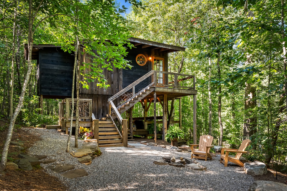 A wooden treehouse nestled among tall trees is accessed by a set of steps. Gravel pathways lead to the entrance, with natural stone accents and outdoor seating present nearby. Green foliage surrounds the structure, creating a secluded atmosphere.