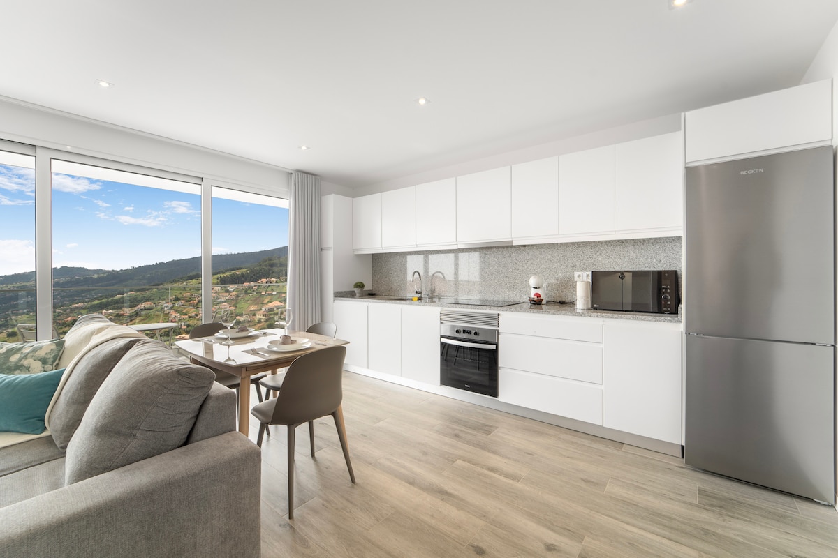 A modern kitchen is displayed with sleek white cabinetry and a stainless-steel refrigerator. A countertop holds a microwave and coffee maker. A comfortable seating area with a sofa is positioned nearby, while large windows offer scenic views of the surrounding landscape.
