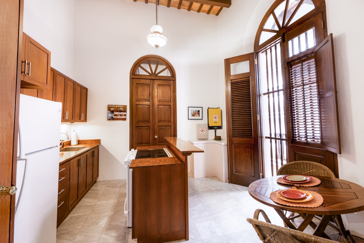 A spacious kitchen area is highlighted by wooden cabinetry and marble countertops. The room features an arched entryway with tall, wooden doors, and a round dining table adorned with decorative plates. Natural light streams through the window with wooden blinds, enhancing the vibrant atmosphere.