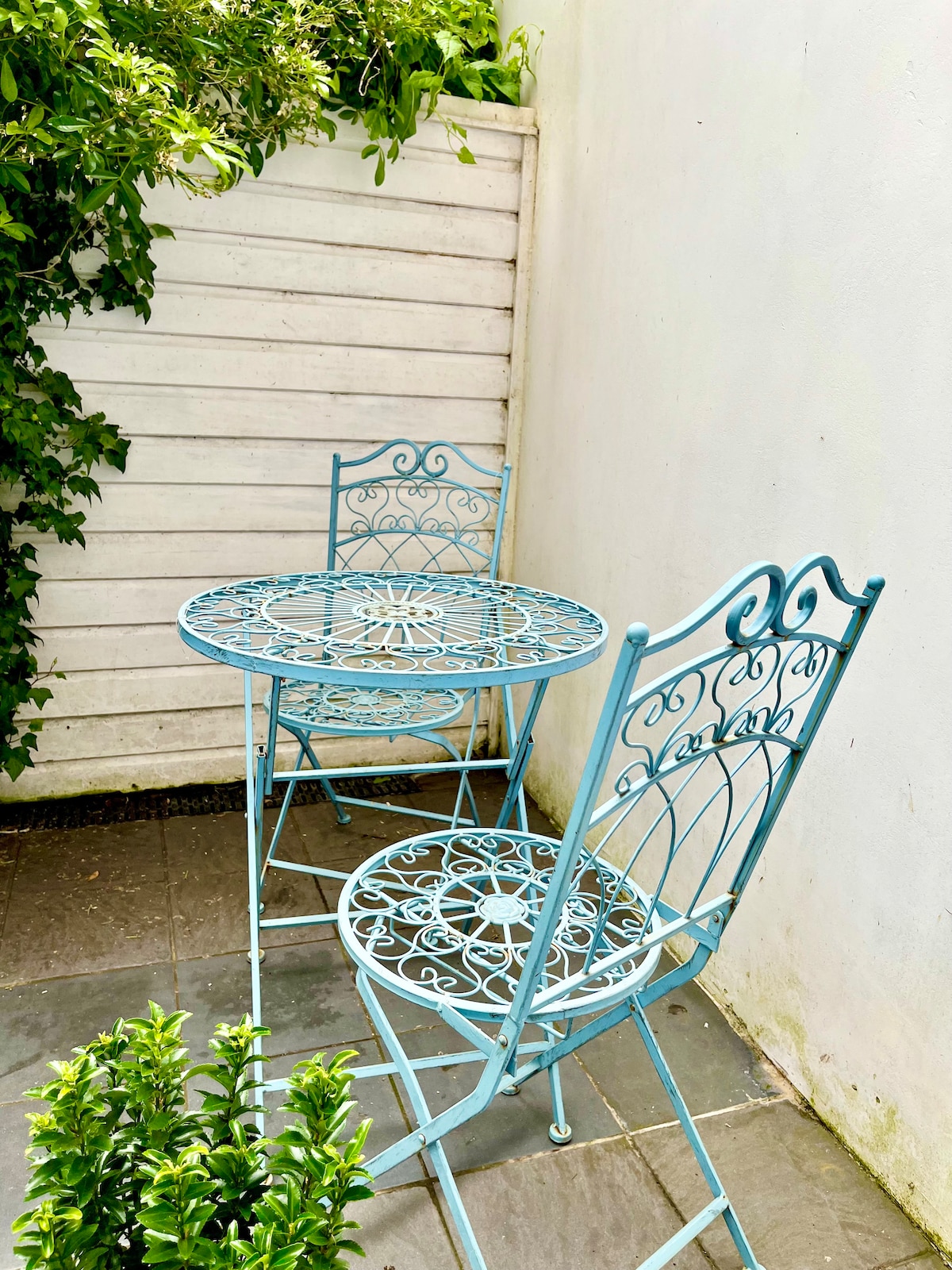 A small courtyard garden is shown, featuring a blue wrought-iron table and two chairs. The furniture displays intricate designs and is set against a white wall, surrounded by green foliage that enhances the outdoor space's charm.