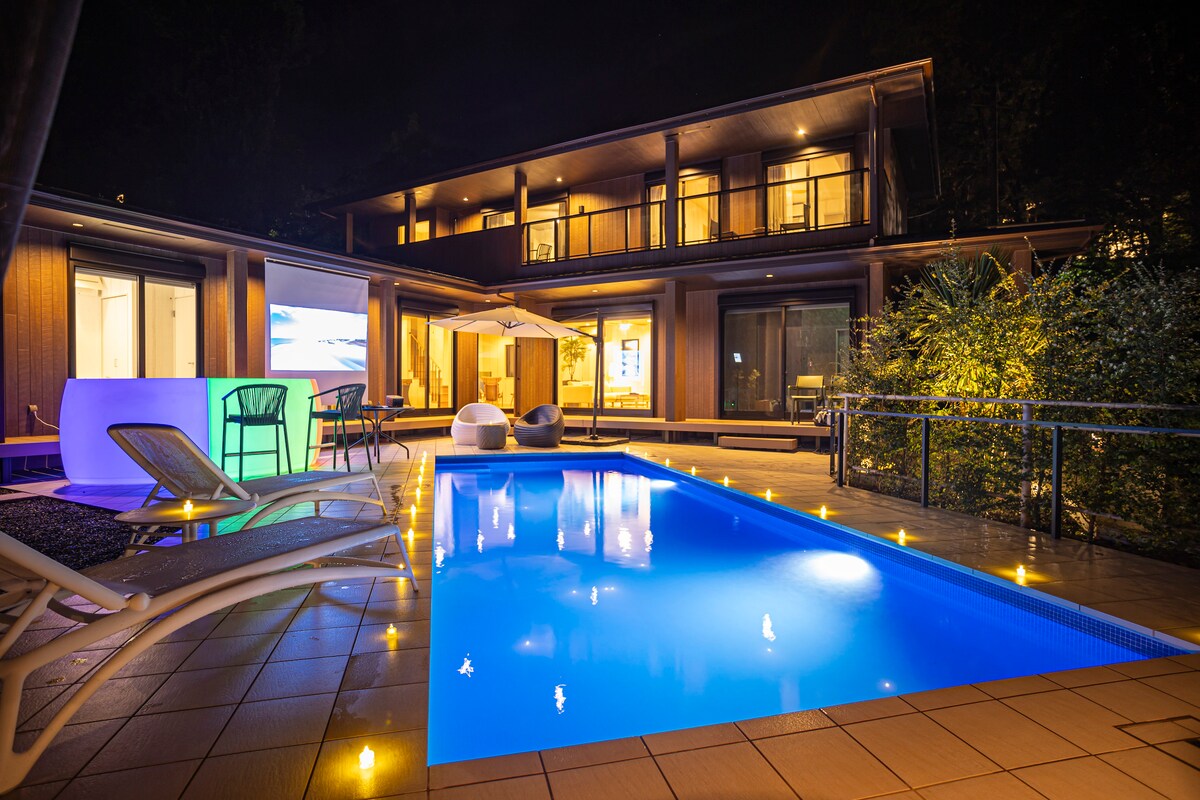 An illuminated pool reflects the deep blue water, surrounded by floating candles that create a serene ambiance. The modern, two-story house features large glass doors, providing a view of the outdoor space. Comfortable lounge chairs and a shaded area enhance the relaxation setting.