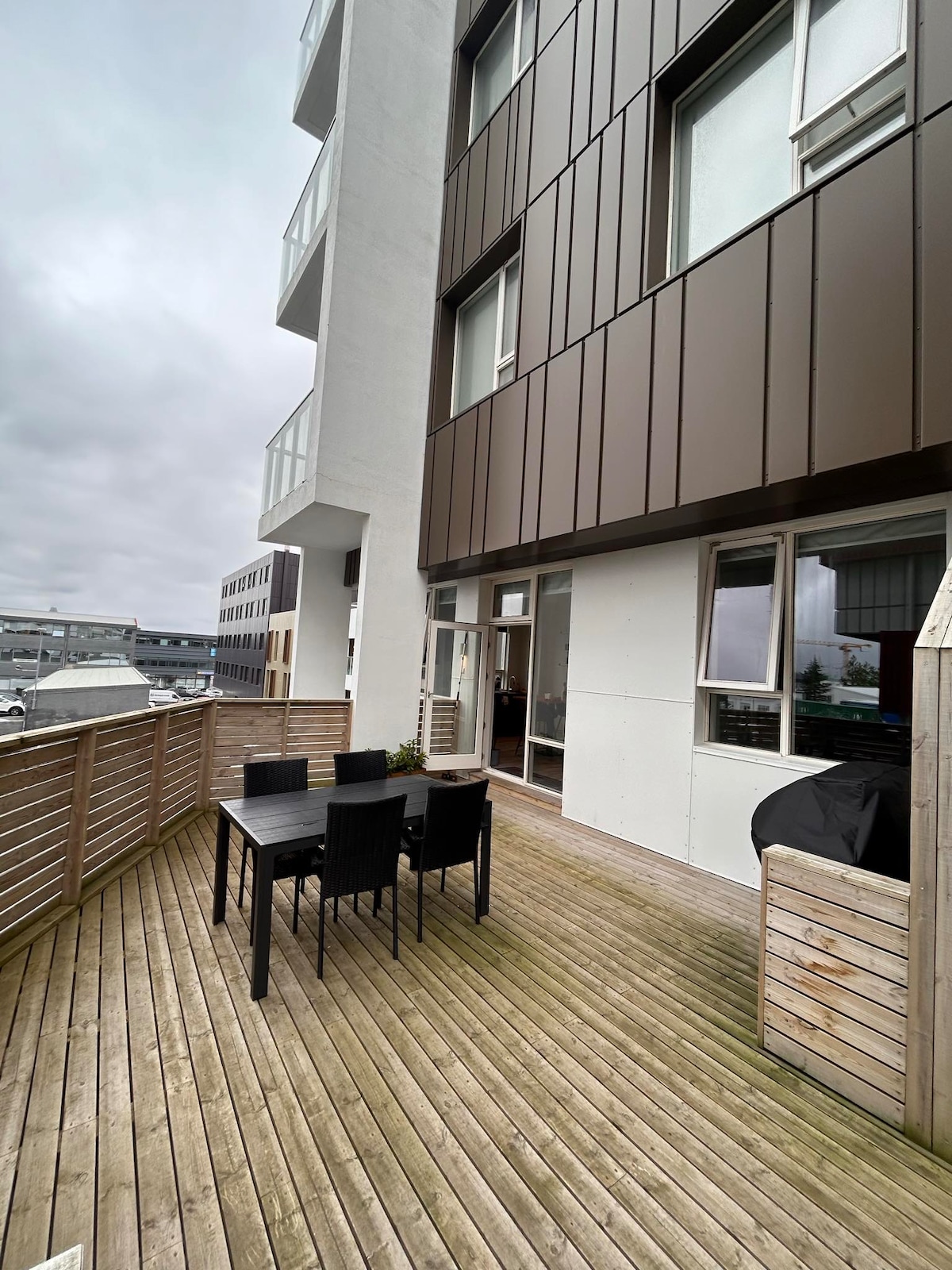 A spacious deck features a wooden floor with a dark dining table surrounded by six black chairs. The exterior of the building showcases a mix of brown and white paneling. Large windows allow natural light into the nearby interior space.