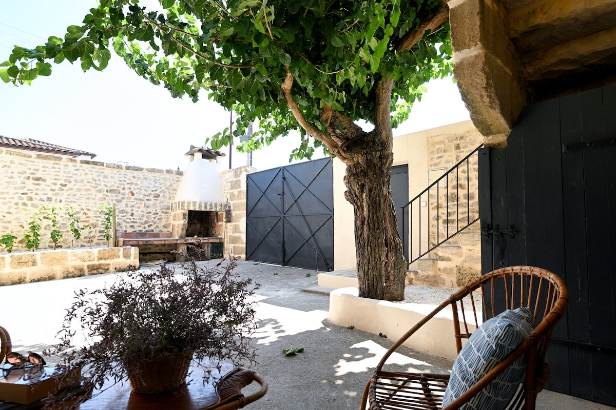 An outdoor courtyard is highlighted by a lush tree providing shade. A rustic barbecue station is visible in the background, and a cozy seating area with a woven chair and decorative pillow invites relaxation. Stone walls frame the space, creating a tranquil atmosphere.