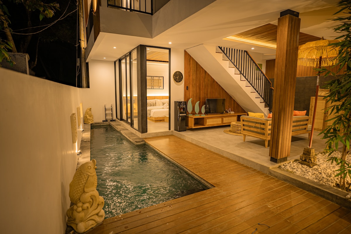 A private pool is featured adjacent to a wooden deck, complemented by decorative stone sculptures. The modern living area, visible through expansive glass doors, is adorned with neutral furnishings. A staircase leads to the upper level, enhancing the villa's spacious design.