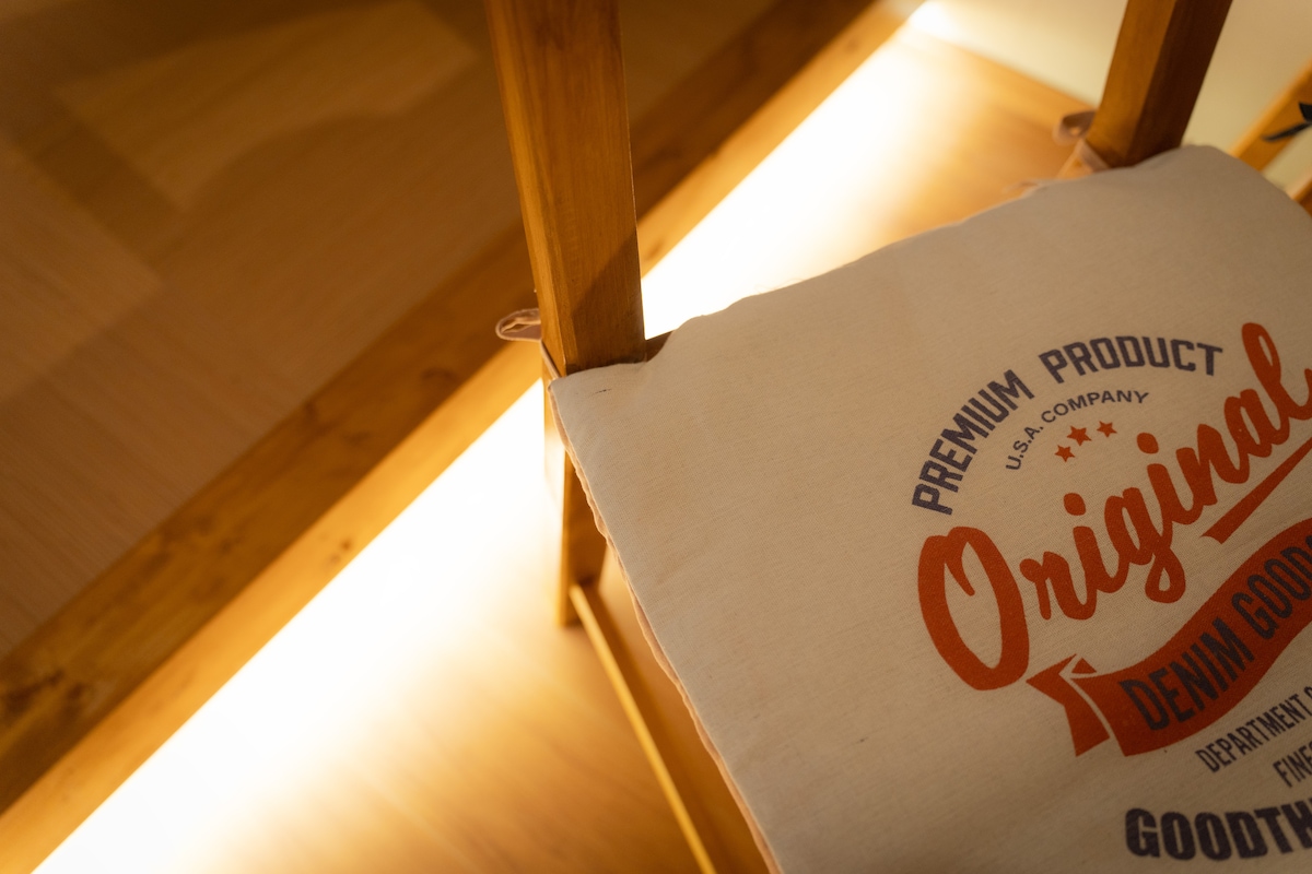 A wooden chair is illuminated softly by a warm light from below. The chair features a cushion with a print indicating 'Premium Product' and 'Original.' The surrounding area conveys a minimalistic aesthetic, emphasizing the natural materials used.