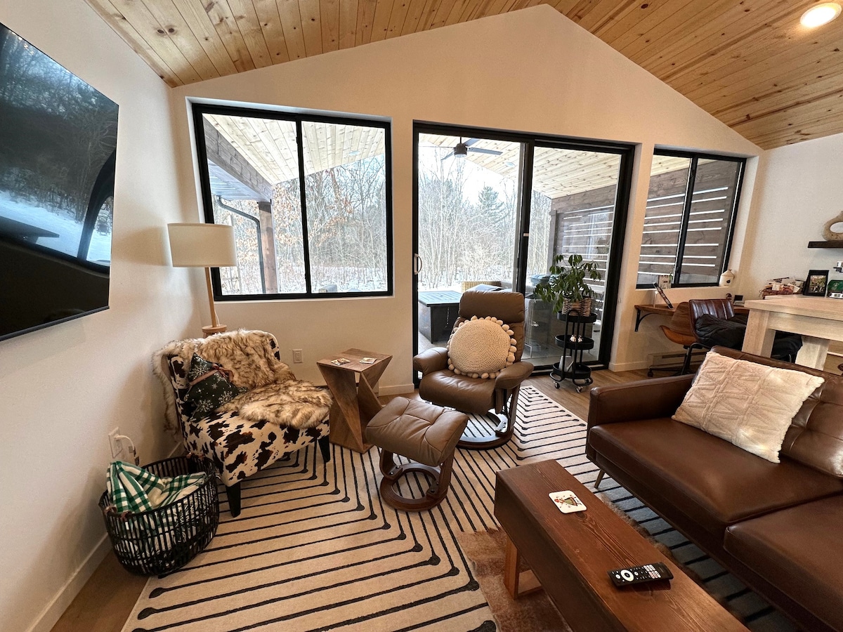 The living room features a mix of seating options including a leather sectional and a cozy accent chair. Large windows invite natural light and offer views of the covered patio. A light-colored area rug and wooden ceiling create a warm, inviting space.