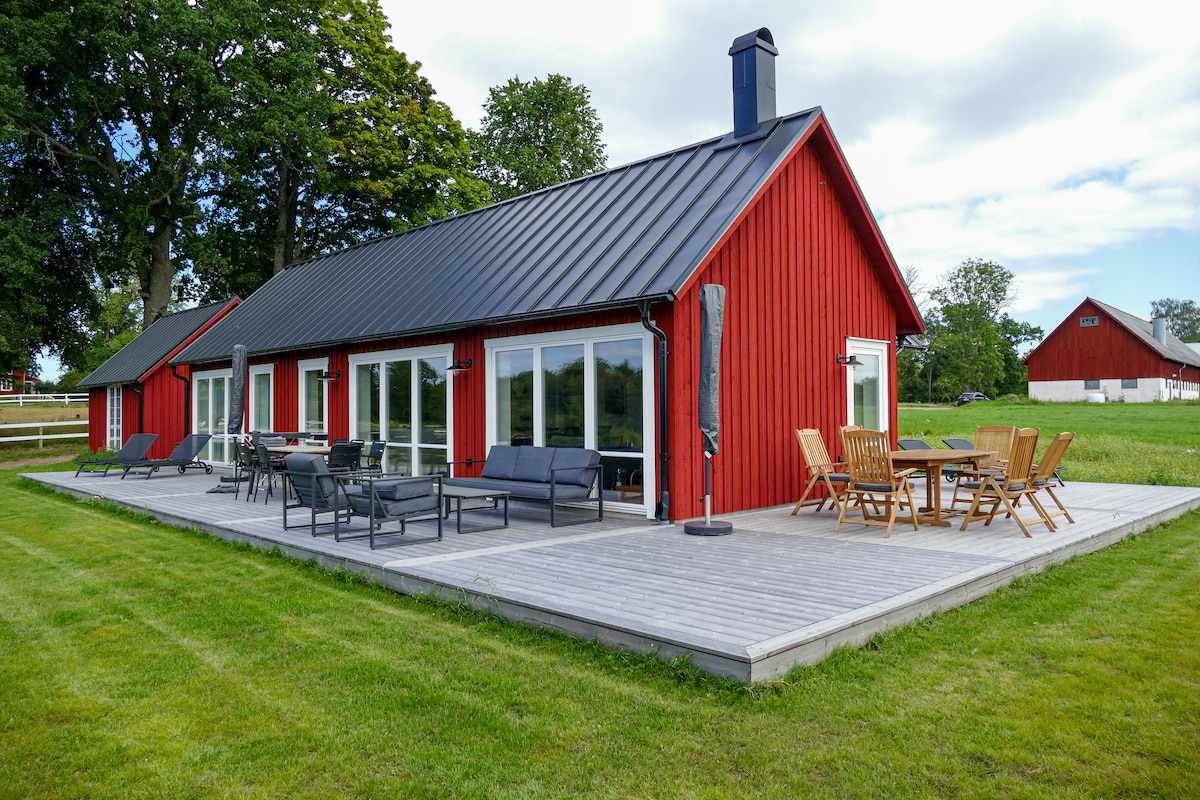 A red wooden house is set against a green landscape, featuring large glass doors that open to a spacious deck. Outdoor seating includes a combination of lounge chairs and a dining table. Nearby, green lawns and trees contribute to the serene environment.