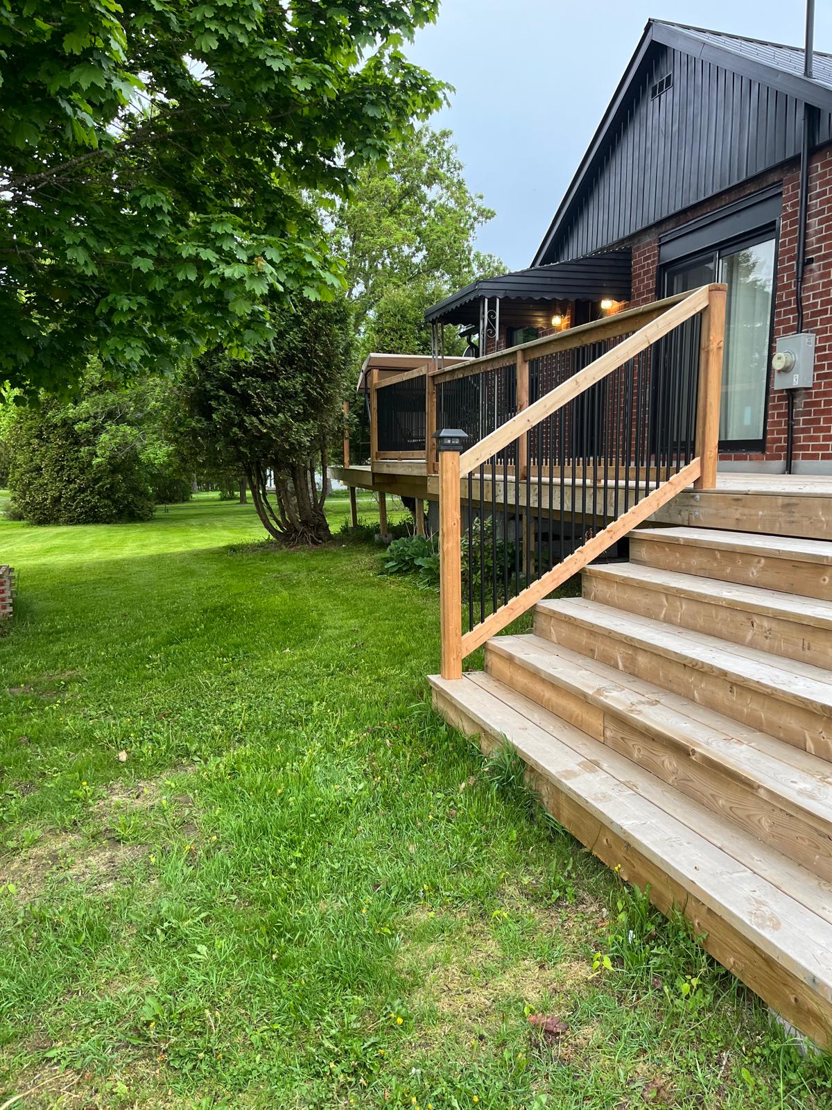 A welcoming entrance features a wooden staircase leading up to the home. A sturdy railing provides safety, while grass and trees create a natural setting in the background, highlighting the peaceful surroundings.