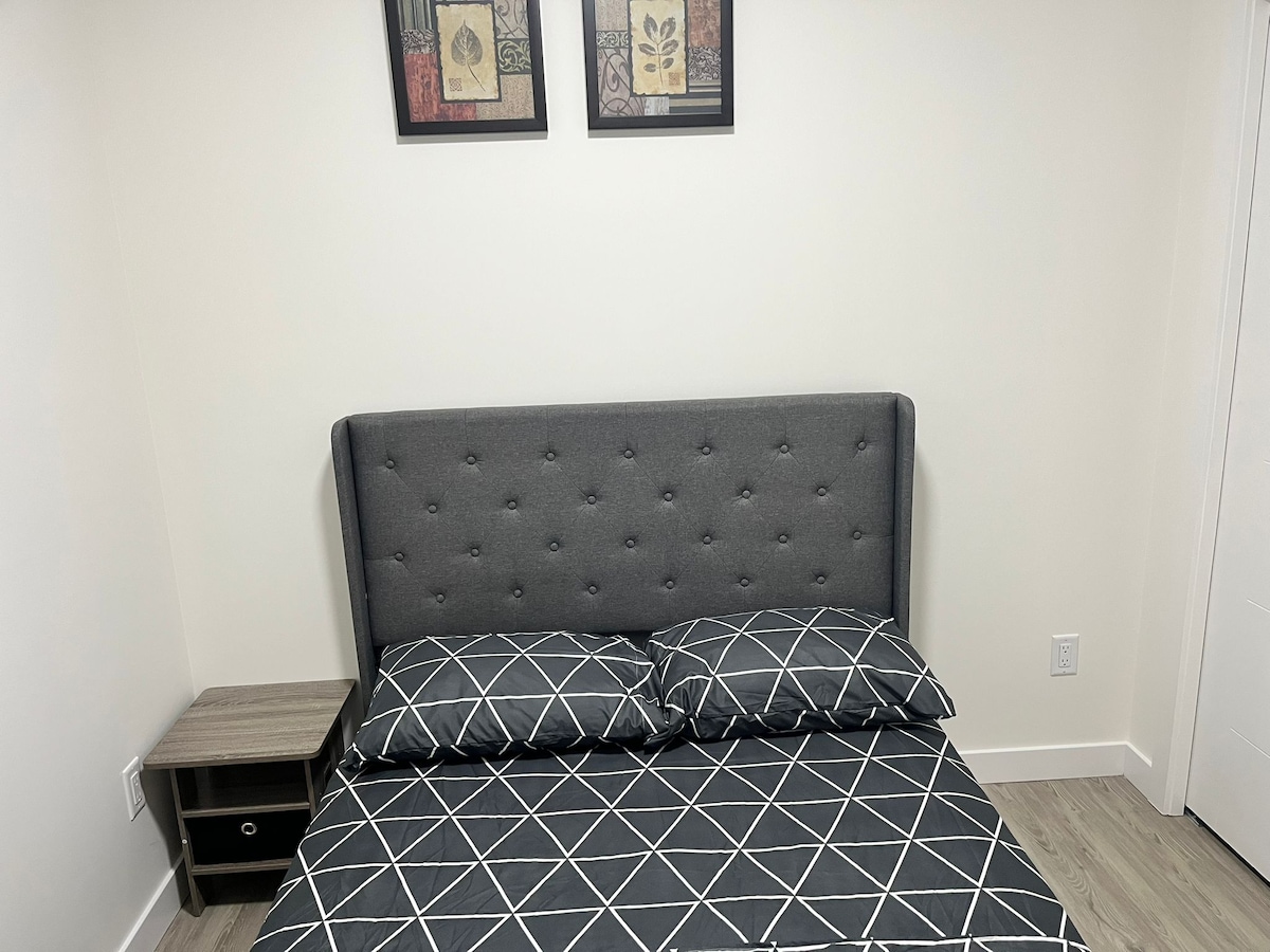 A cozy bedroom features a gray upholstered headboard with subtle tufting. A patterned duvet cover with geometric designs rests on the spacious bed, complemented by two matching pillows. Framed artwork hangs above, while a small nightstand is situated beside the bed.