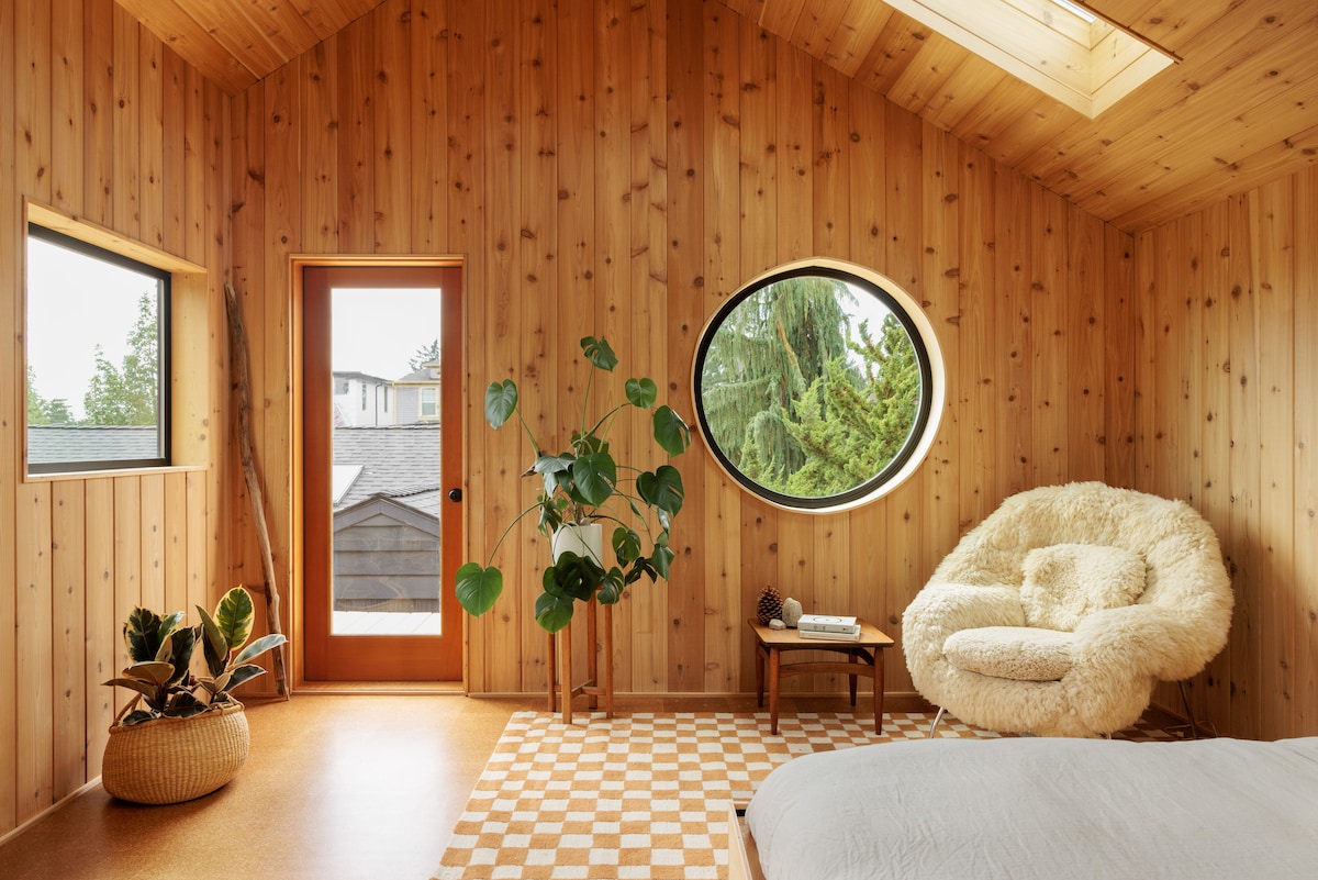 An interior view reveals warm wooden walls and a vaulted ceiling. A round window frames greenery outside. A cozy chair and a plant add a touch of nature, while a patterned rug anchors the space near a bed and a door leading to the exterior.