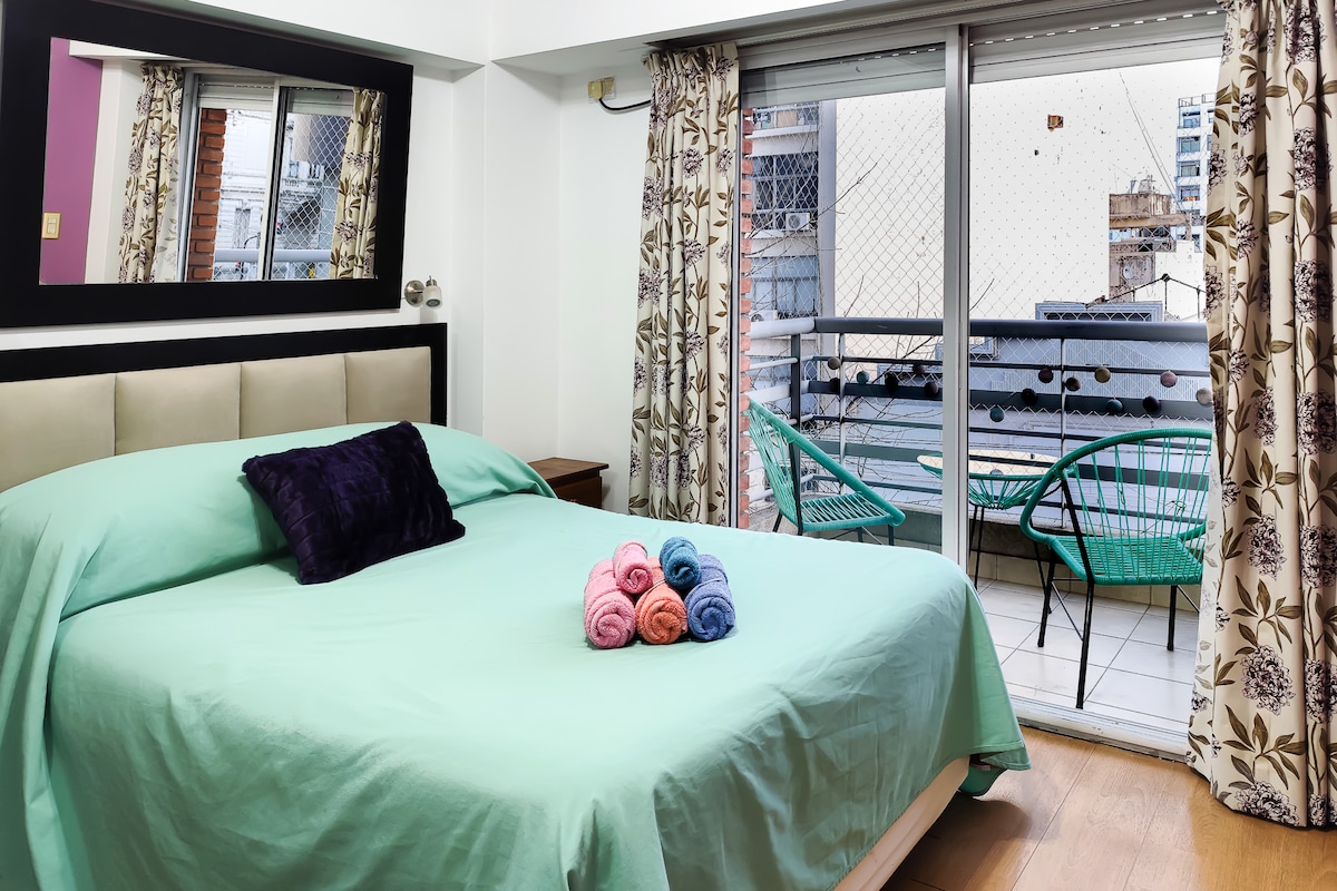 A spacious bed with a light green coverlet is featured, accented by a dark pillow. Colorful rolled towels are arranged on the bed. A large window opens to a balcony, which is furnished with green chairs and adorned with floral curtains.