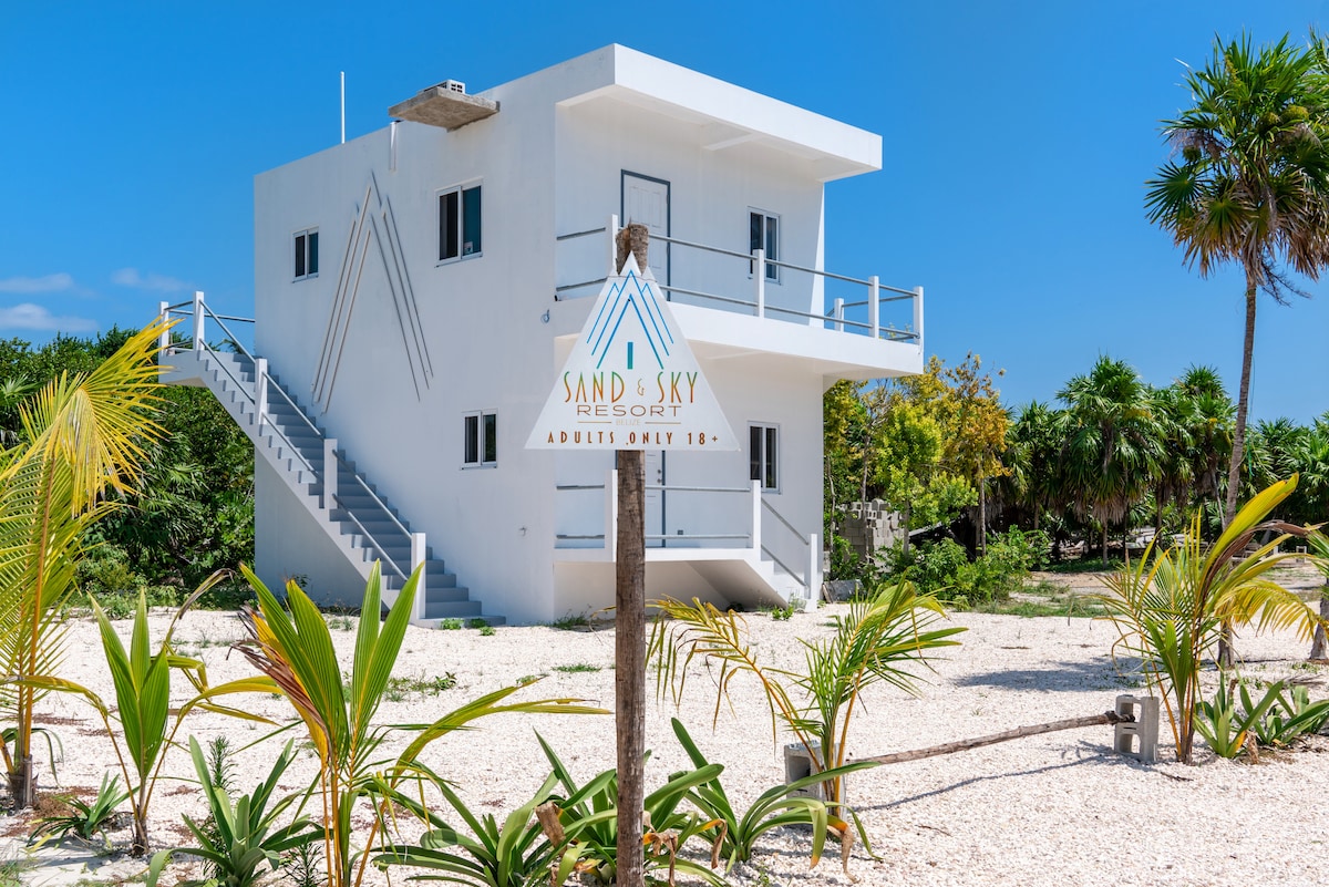 A modern two-story A-frame resort structure is framed by tropical greenery and white pebbles. A clear blue sky adds to the bright atmosphere, while a wooden post displays the resort's logo. A staircase leads to the upper level, providing access to elevated views.