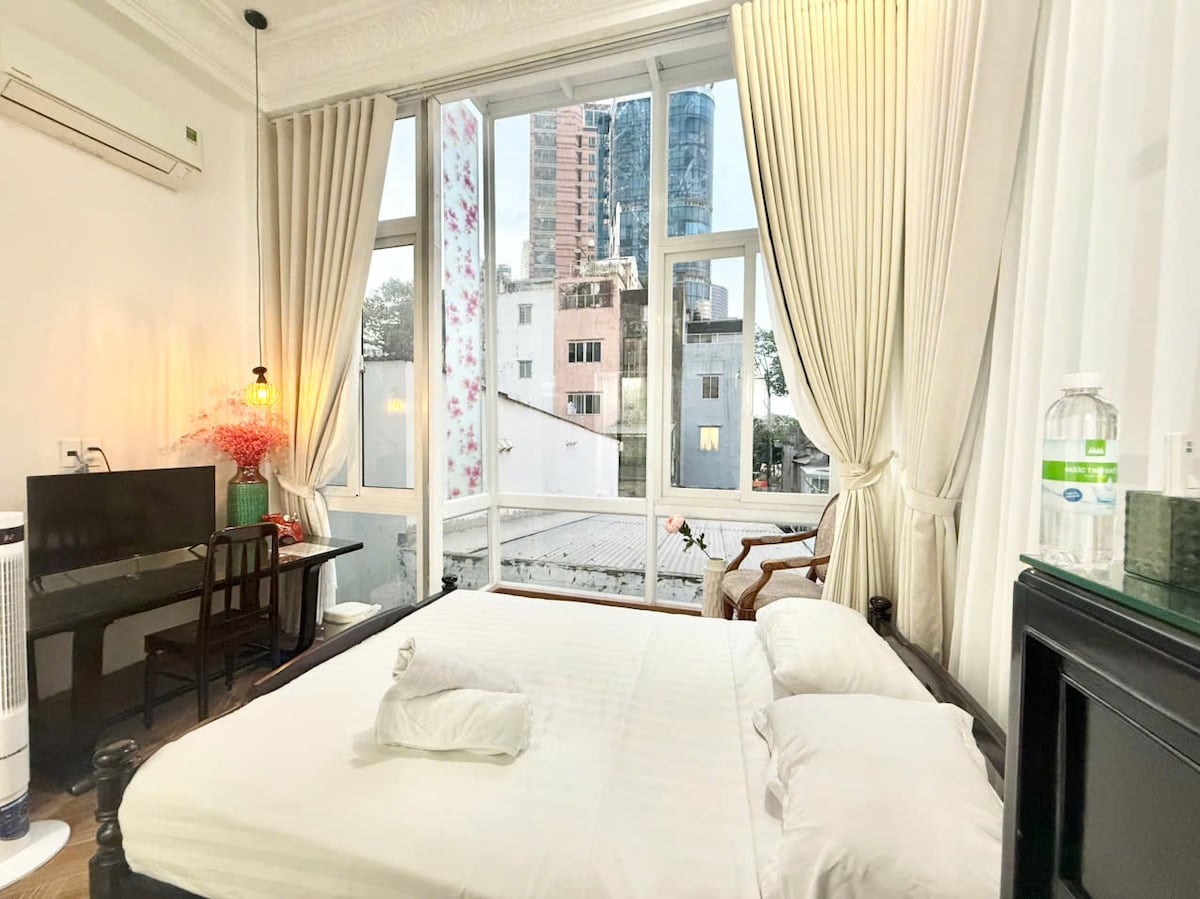 A bright sleeping area includes a spacious bed with a single pillow. Large windows are draped in soft curtains, allowing natural light to fill the room. A small desk and chair are positioned near the window, with a decorative plant adding a touch of color.