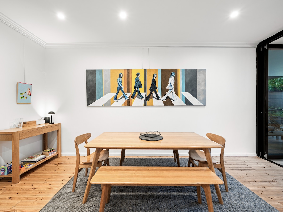 A dining area features a wooden dining table surrounded by matching benches and chairs. An abstract artwork is displayed on the wall above, contrasting with the white walls. Natural light from large glass doors illuminates the space, enhancing the modern and inviting atmosphere.