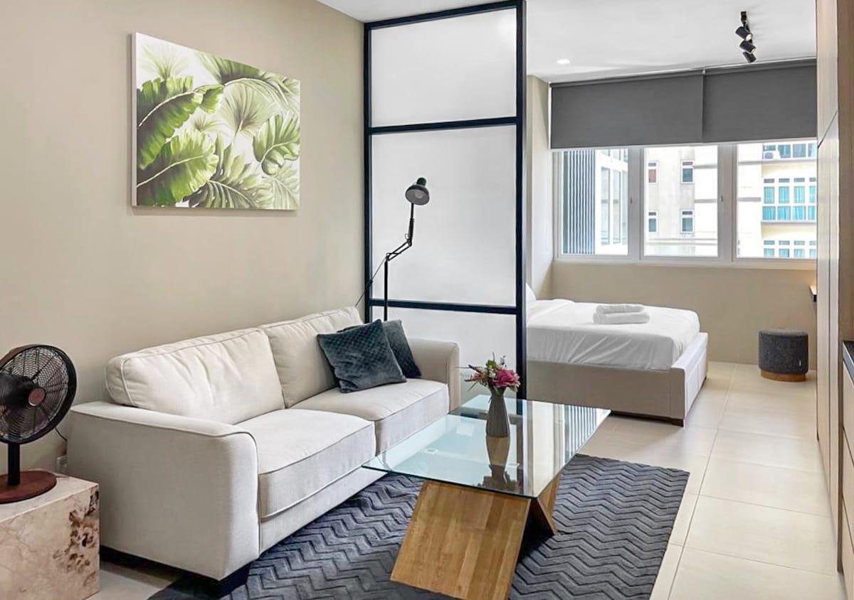 The open living area features a light-colored sofa accompanied by a glass coffee table and a standing lamp. A decorative wall art piece with green leaves adds a touch of nature. The bedroom area is partially separated by a frosted glass partition and includes a queen-sized bed with white linens.