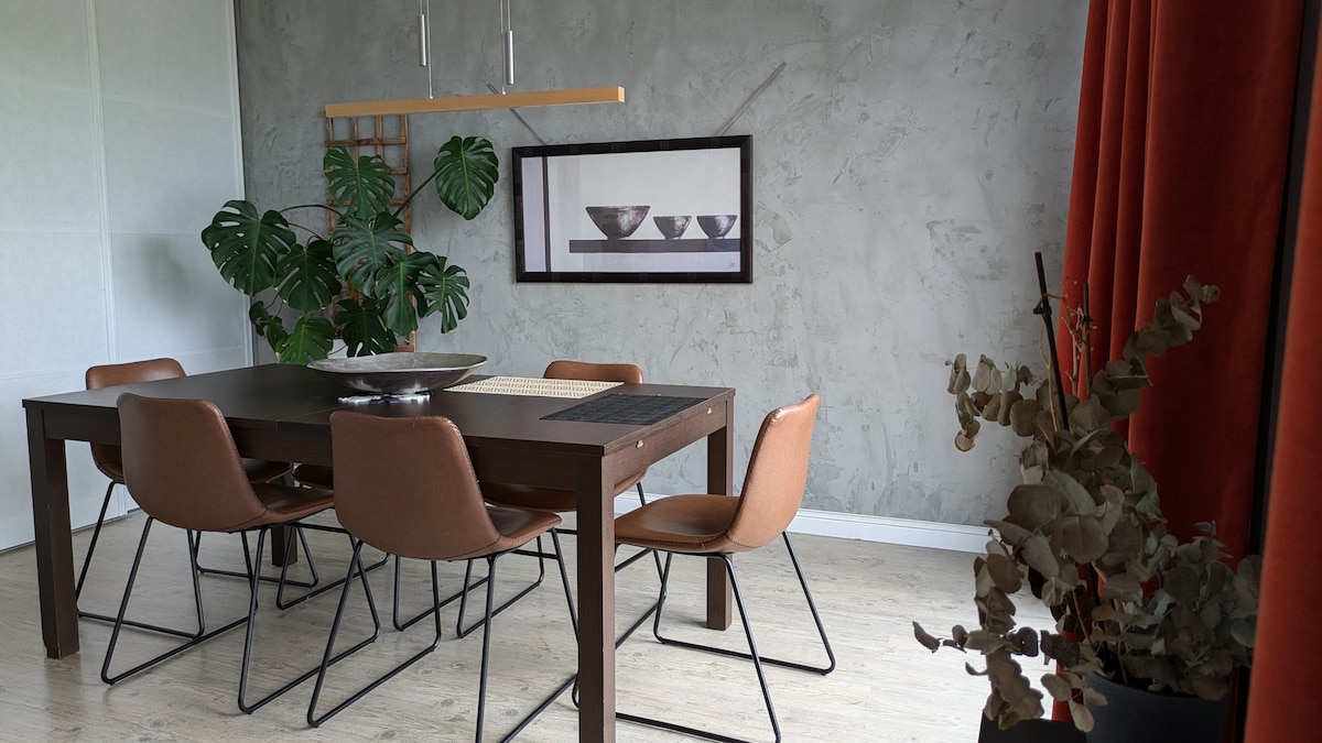A spacious dining area features a dark wooden table surrounded by six brown chairs. A minimalistic artwork depicting bowls is displayed on the wall. Green plants are positioned near the window, and textured grey walls complement the warm orange curtains.