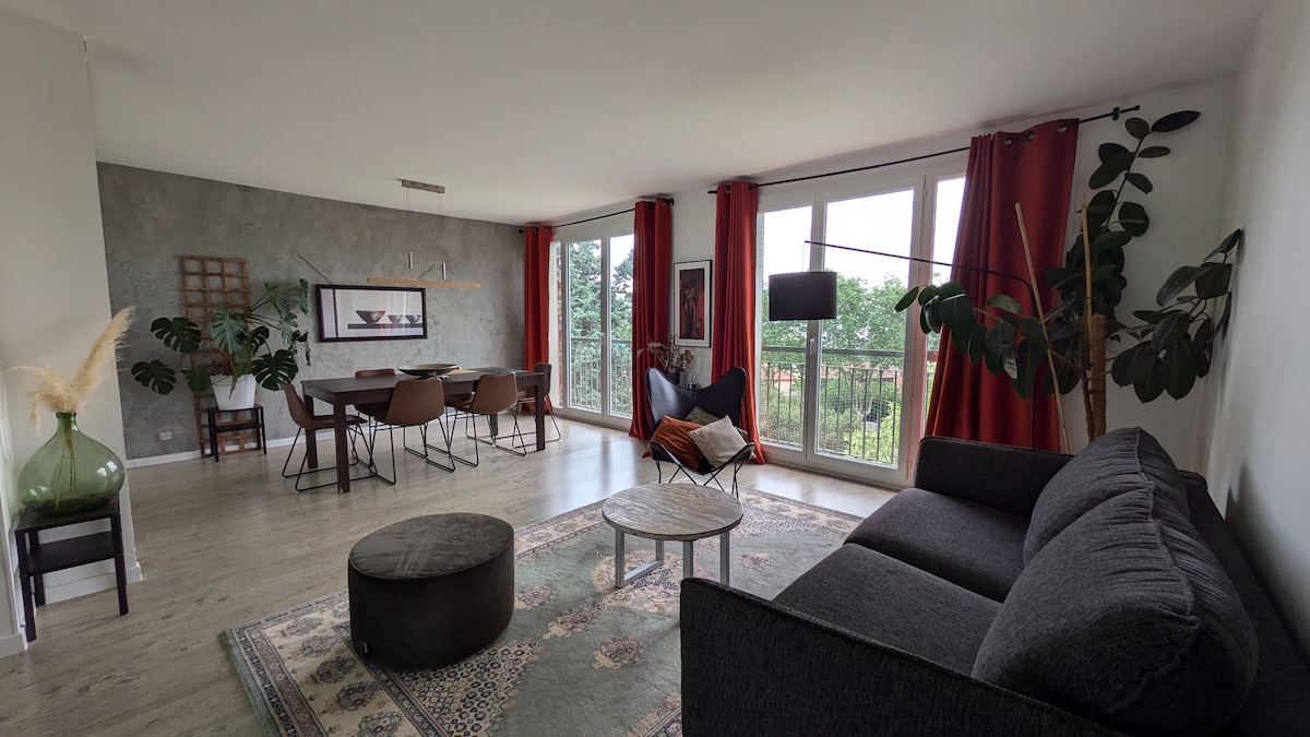 A spacious living area features large windows with red curtains, allowing natural light to fill the room. A comfortable sofa and decorative plants enhance the space, while a dining table for six provides ample seating. A round coffee table is positioned on a patterned rug.