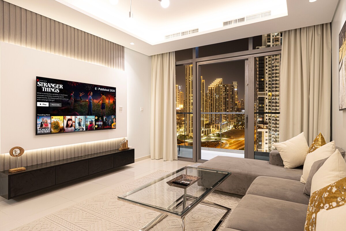 A modern living room is presented, featuring a plush L-sofa adorned with decorative pillows. A 65" Smart TV is mounted on the wall, showcasing a media interface. Beneath the TV, a sleek glass coffee table complements the contemporary décor, with city lights visible through large windows.